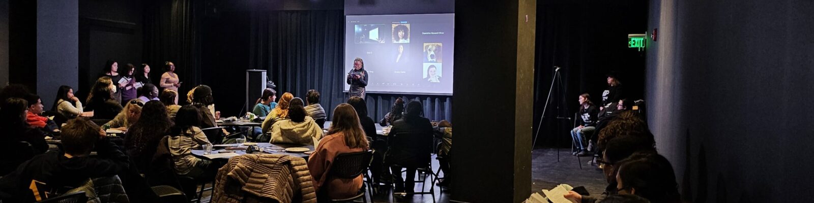 Image of a student reading at a microphone to a room full of people as well as Zoom attendants