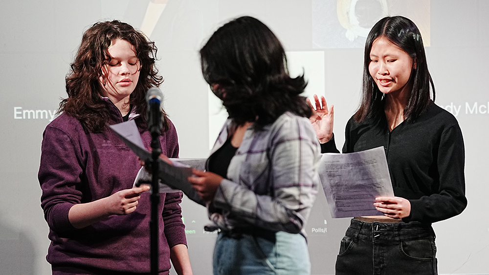 Three students standing near a microphone before a stage performance