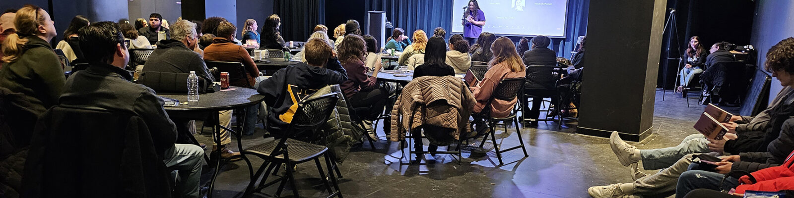 An audience in chairs, seated in front of a stage for a reading performance