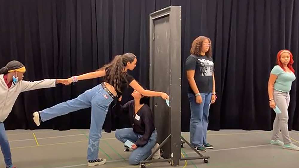 A group of Emerson students participating in a theatre performance or exercise held in a practice studio; multiple students are standing in various postures behind and in front of a doorway